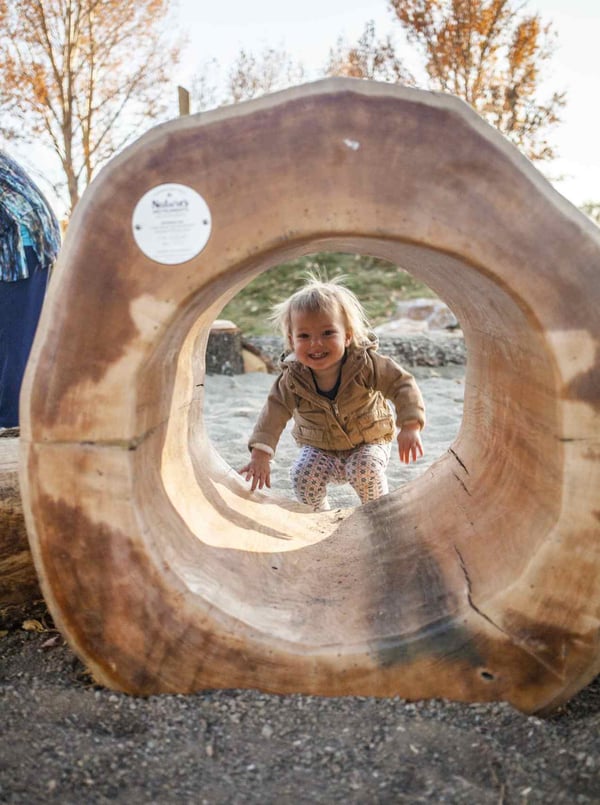 A toddler peeks through a log at a natural playground at Rio Grande Farm Park